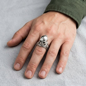 Closed silver skull locket ring worn on a hand resting on light grey canvas.