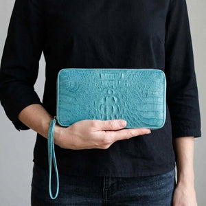 Cartera con cremallera de piel de cocodrilo hornback azul cielo, sostenida con naturalidad por una mujer adulta