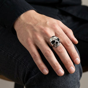 Sterling silver skull ring worn on a relaxed hand resting on a dark denim-covered knee.
