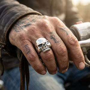 Close-up of a rugged, tattooed hand gripping a motorcycle handlebar while wearing a polished sterling silver skull ring featu