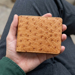 Tan full-quill ostrich bifold supported in a hand, showing its natural raised pattern and matching stitched border.