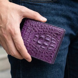 Billetera bifold de piel hornback de cocodrilo violeta sostenida junto a tela vaquera oscura