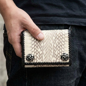 White cobra leather biker wallet held at the top edge, showing cream scales, black braided trim and two silver-tone corner ornaments